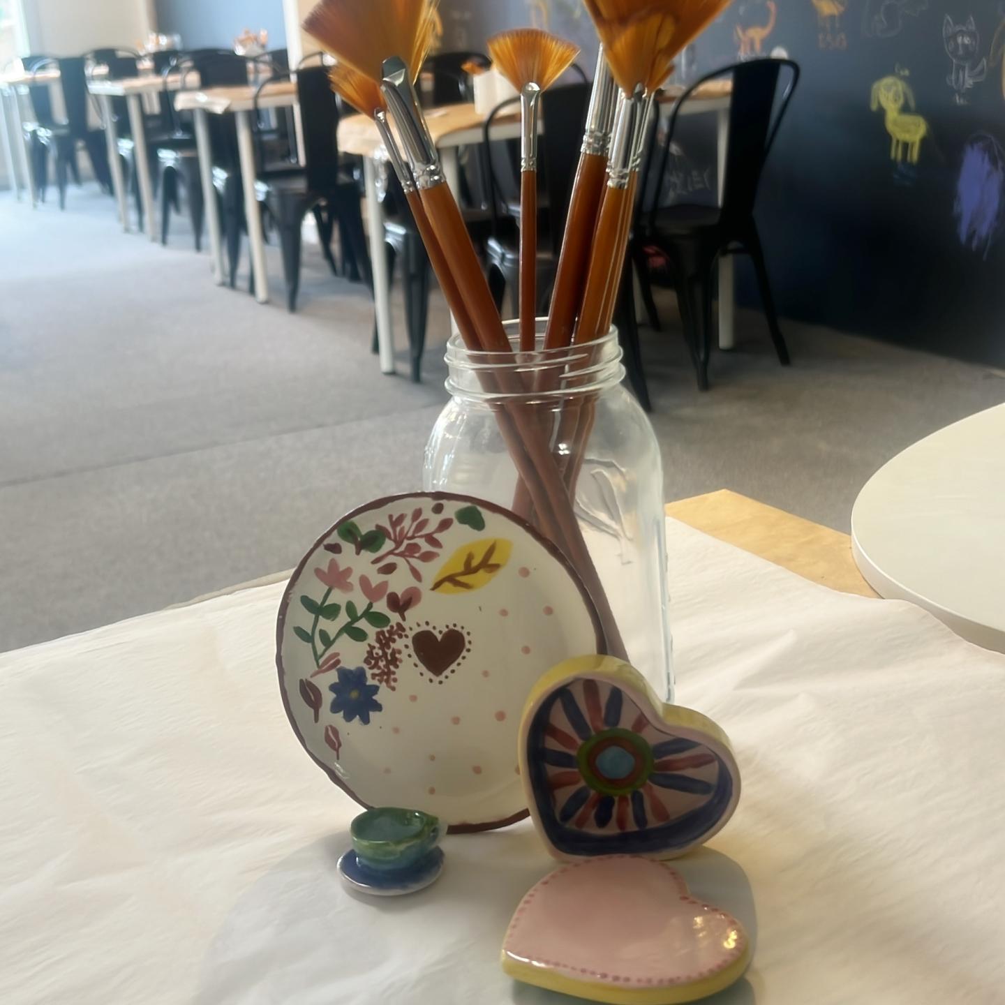 Paintbrushes in a jar with painted ceramic disks and hearts on a table.