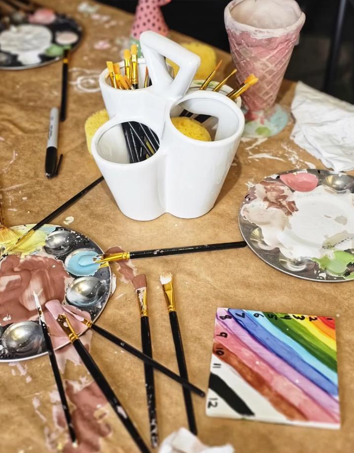 Art supplies on a table: brushes, paint, palettes, and a rainbow gradient paper.