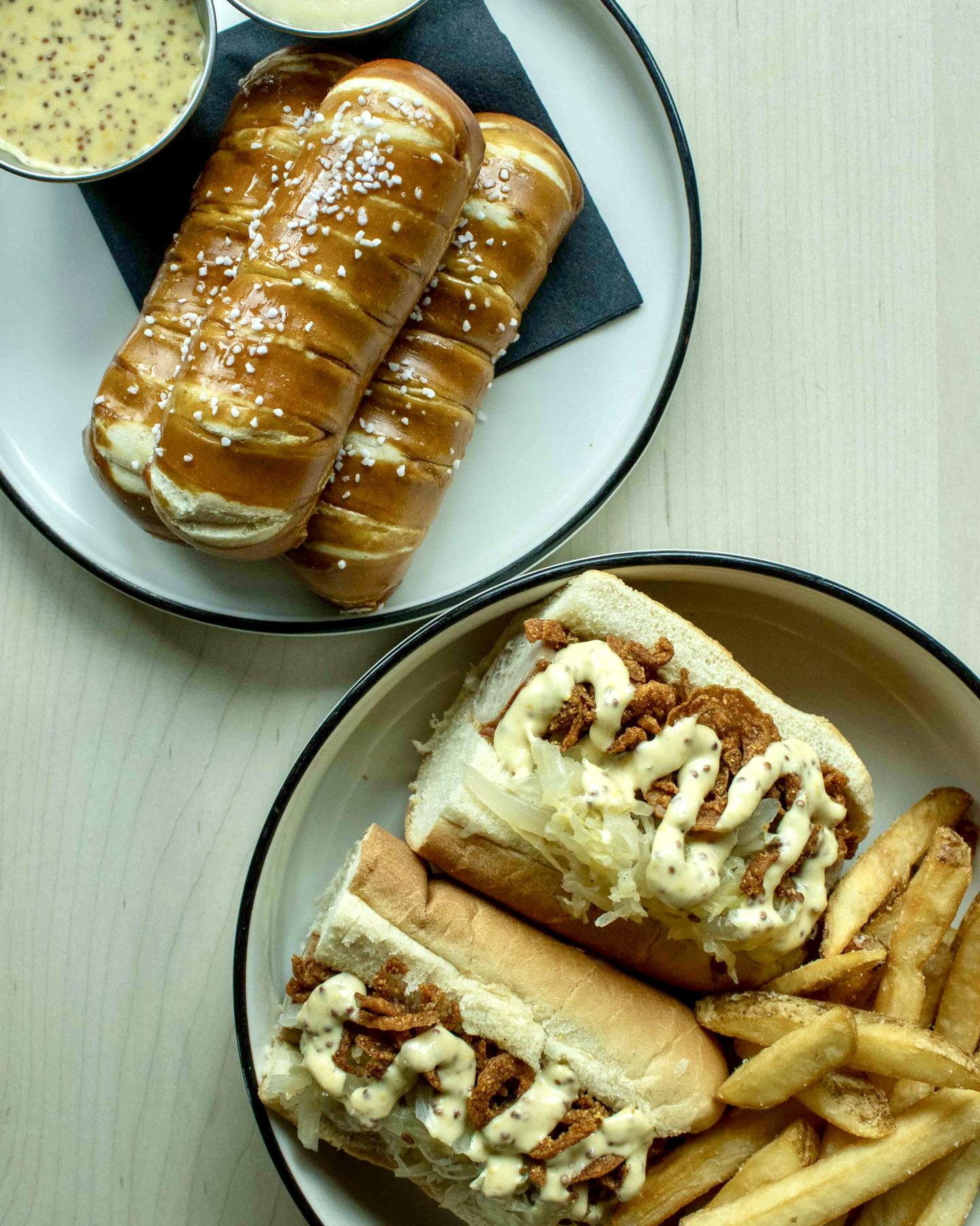 bratwurst sandwiches with fries and two pretzels with mustard dip on plates.