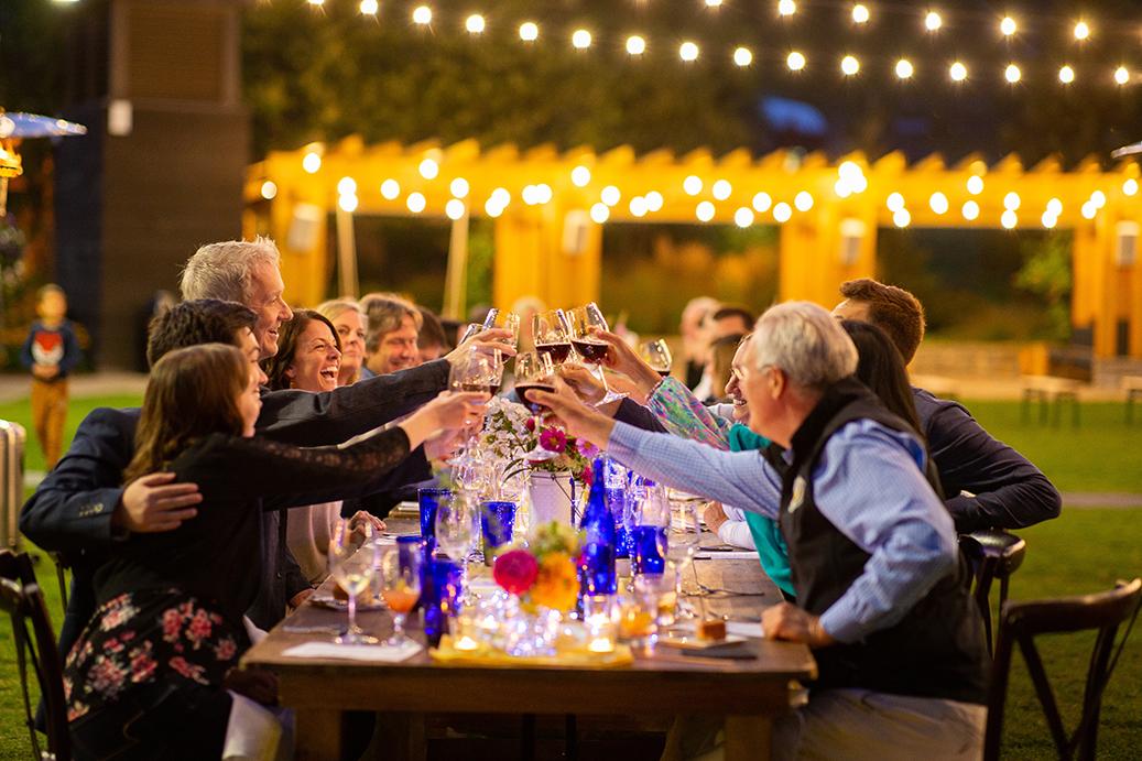 Outdoor dinner party; guests toast under string lights at a long table.