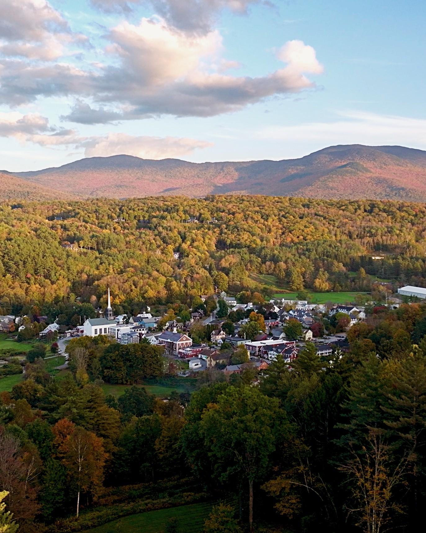 Small town amid autumn trees with mountains in the background under a cloudy sky.