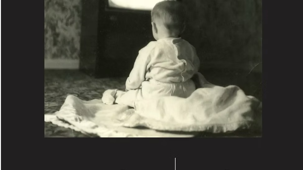 Baby sitting on blanket, watching an old TV in a dim room.