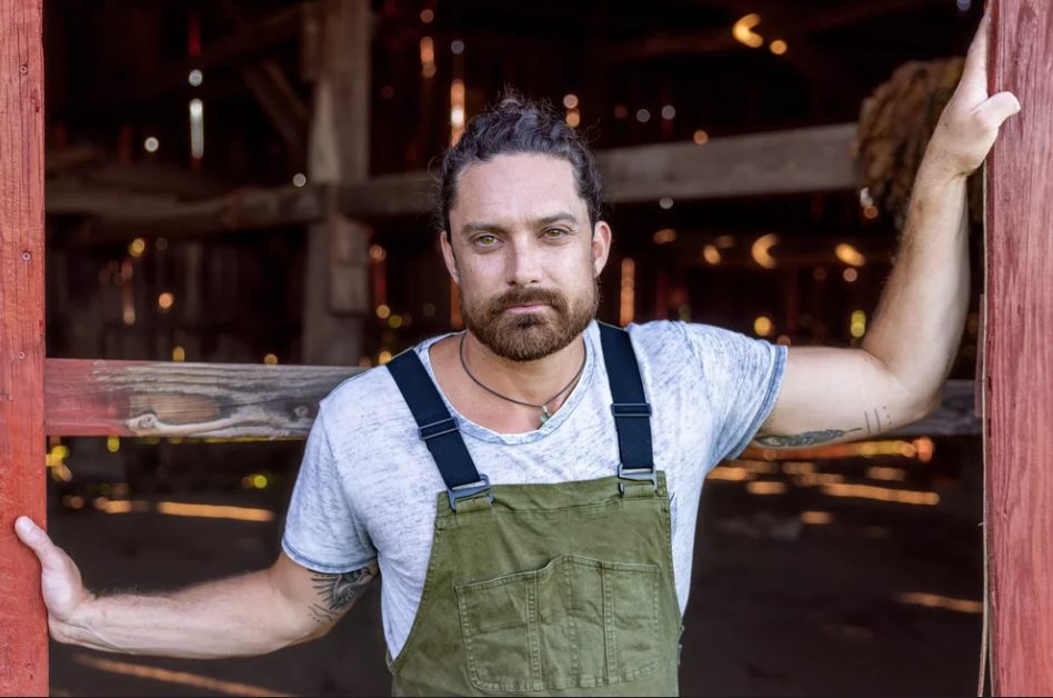 Man in overalls stands in a barn doorway, looking at the camera.
