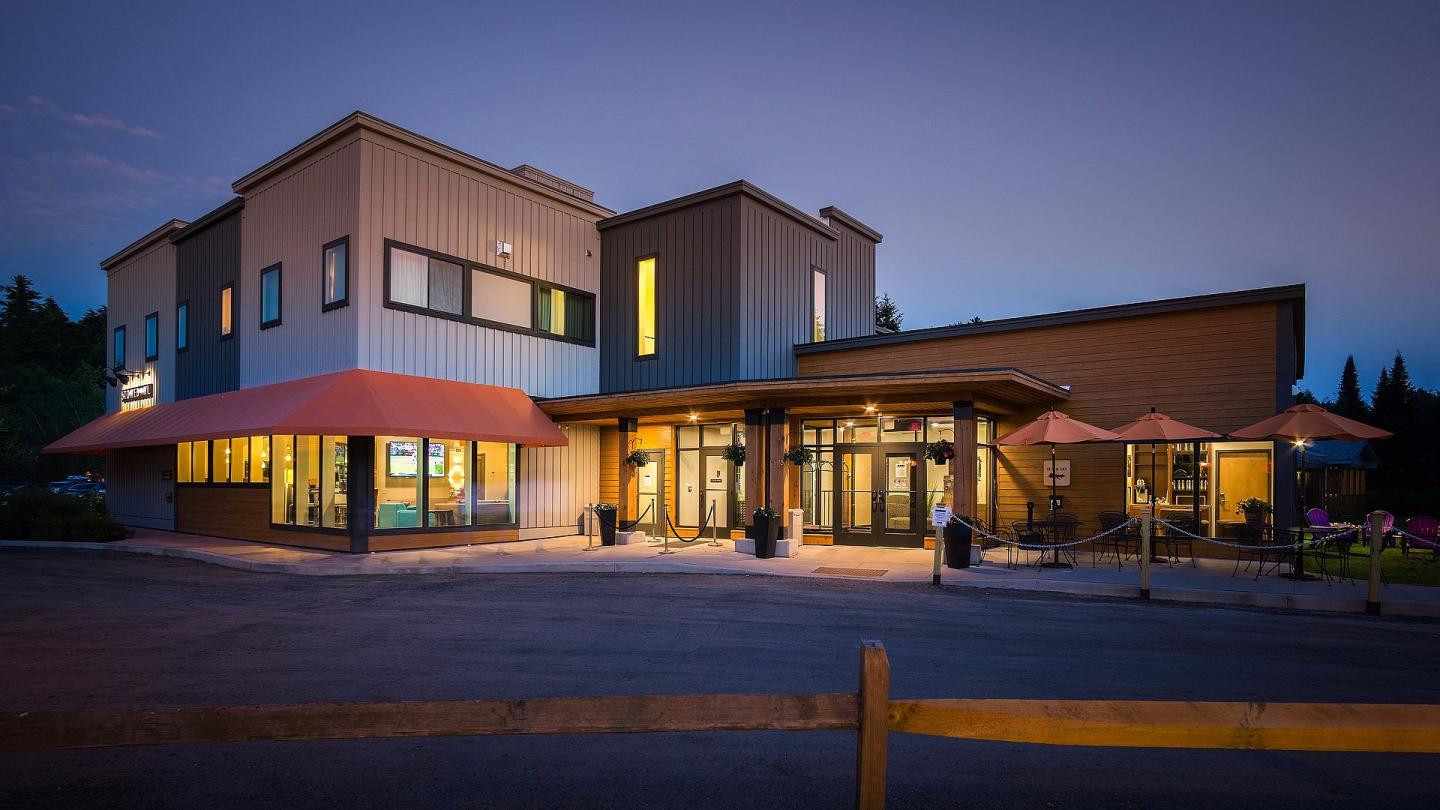 Modern building with warm lighting at dusk, surrounded by outdoor tables with umbrellas.