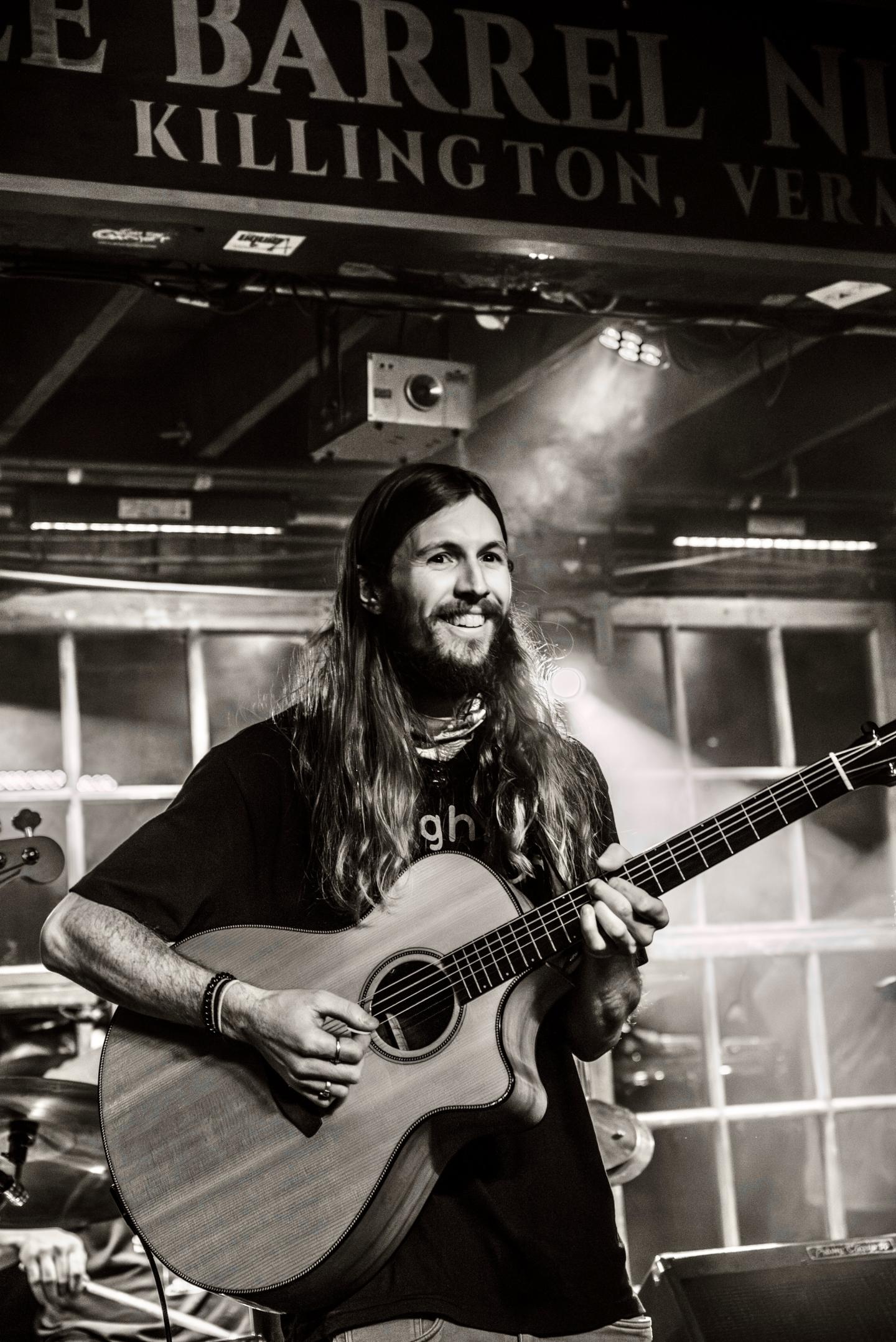 Musician with long hair playing acoustic guitar on stage, smiling. Black and white photo.