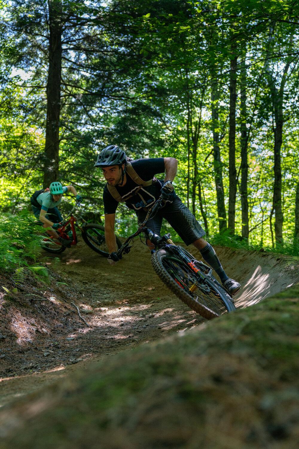 Cyclists racing down a forest trail, surrounded by lush greenery.