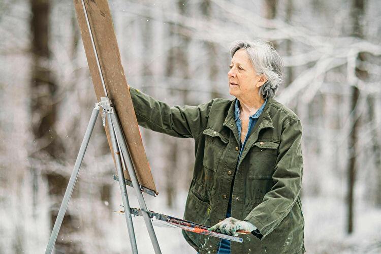 Elderly person painting on canvas in snowy forest.