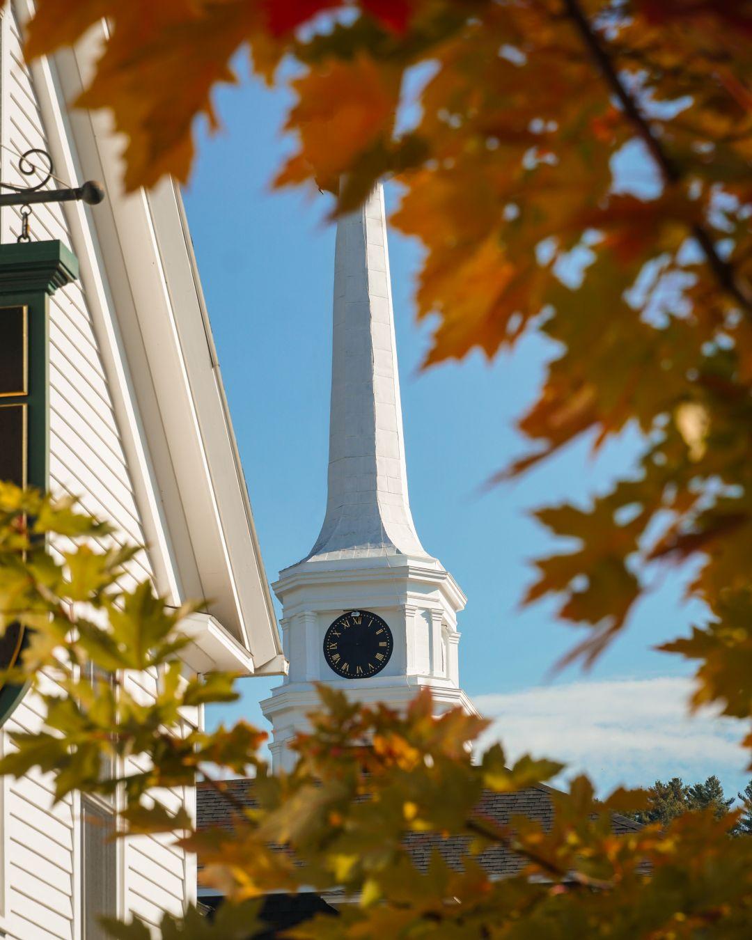 White church steeple with clock, framed by autumn leaves and blue sky.