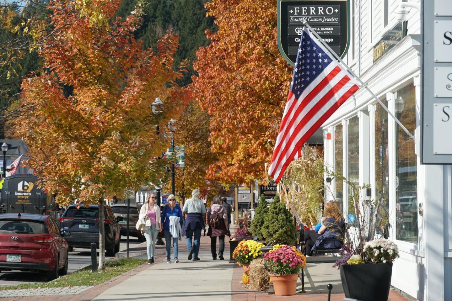 Sidewalk with American flag and autumn trees, people walking nearby.