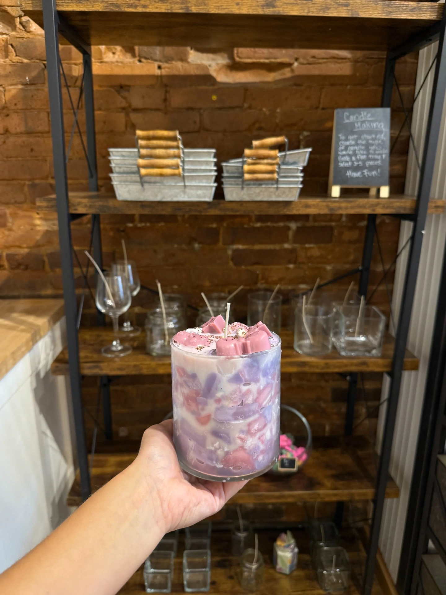 Hand holding a pink and white marbled candle in front of a rustic shelf.