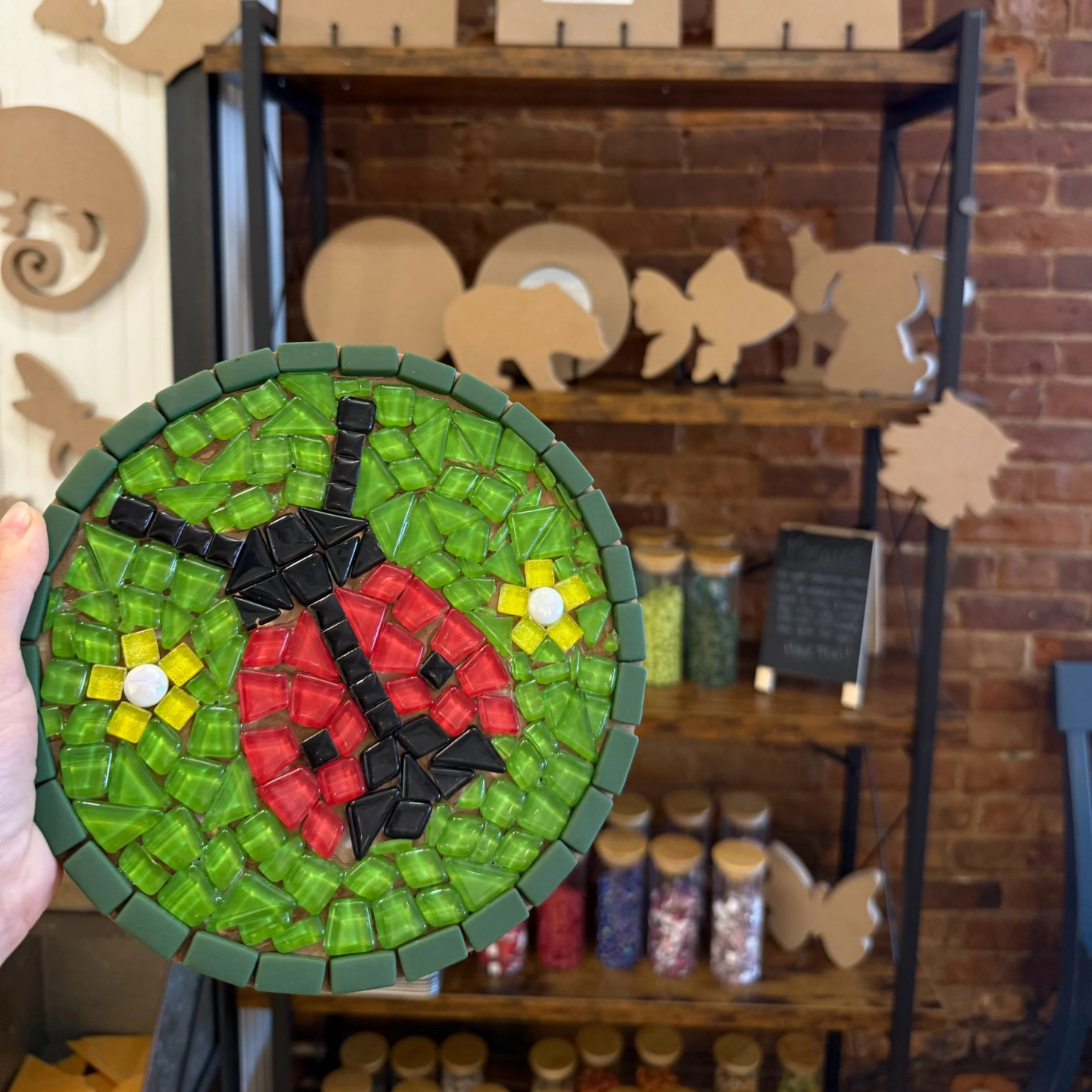 Ladybug mosaic art with green background held in hand, crafting supplies in the background.