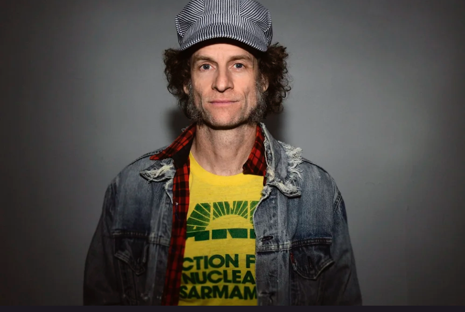 Man wearing a denim jacket and cap with a yellow shirt, standing against a gray background.