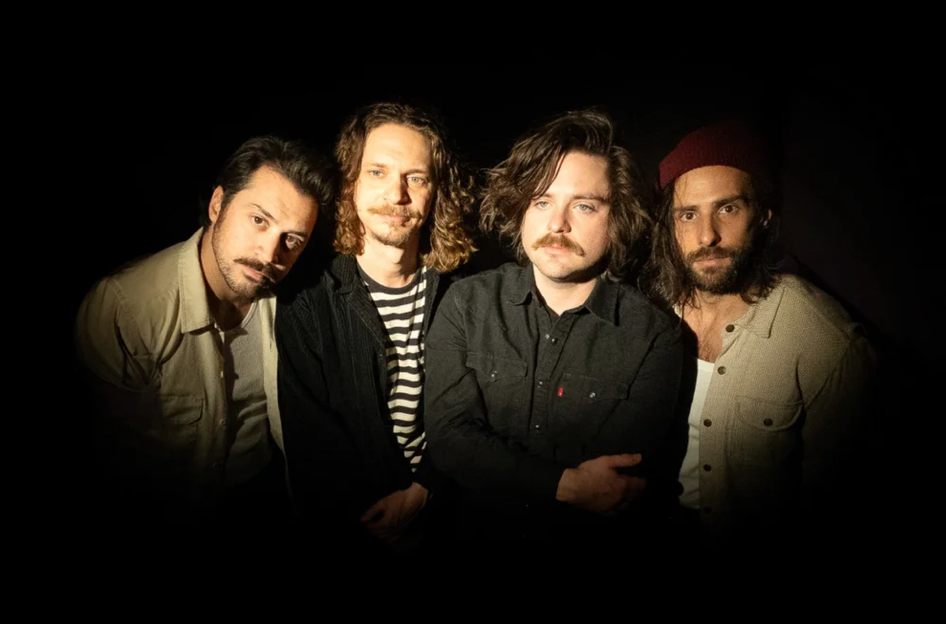 Four men standing closely together in dim lighting, one wearing a red beanie.