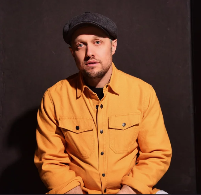 Man in yellow jacket and flat cap against dark background.