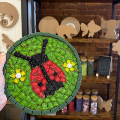 Handcrafted mosaic of a ladybug on green background, shelves with crafts in the background.