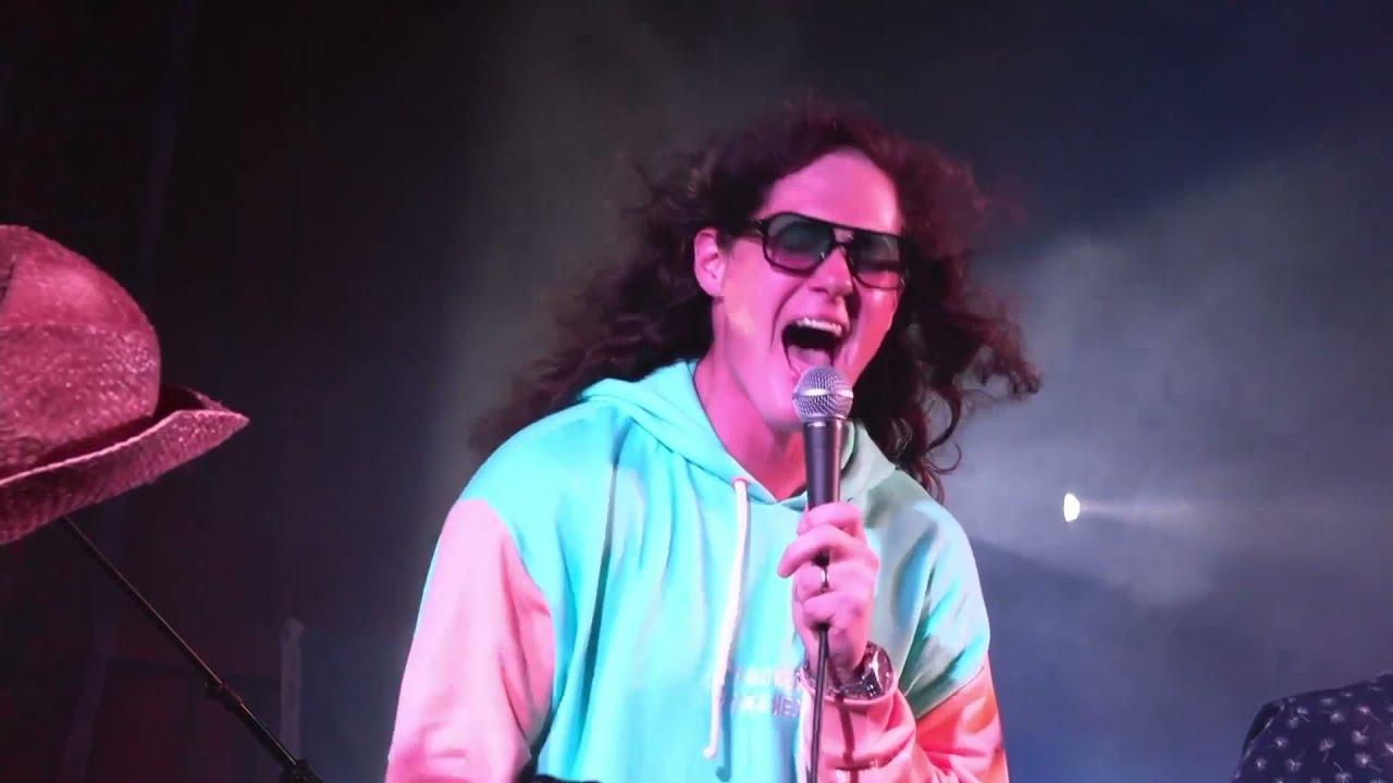 Singer in colorful hoodie and sunglasses energetically performing on stage.