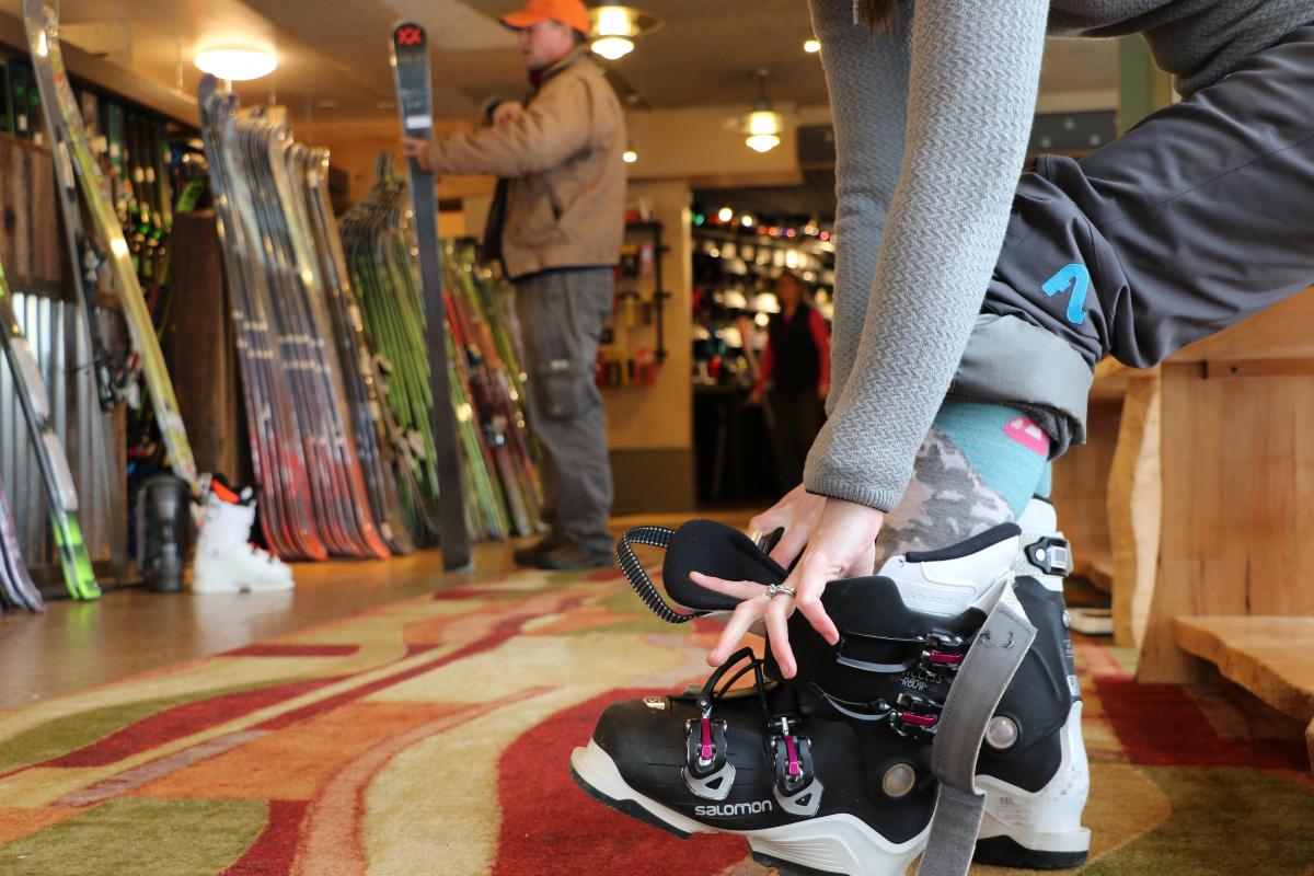Person fastening ski boots in a ski shop, with skis and equipment in the background.