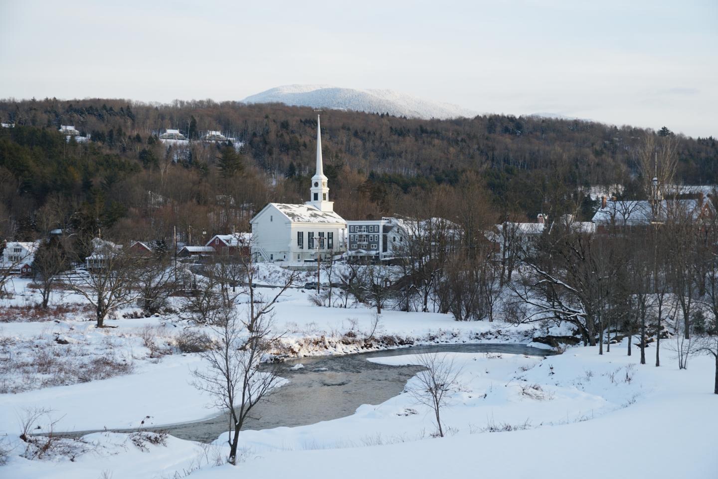 Snowy village with a white church, river, and distant hills.