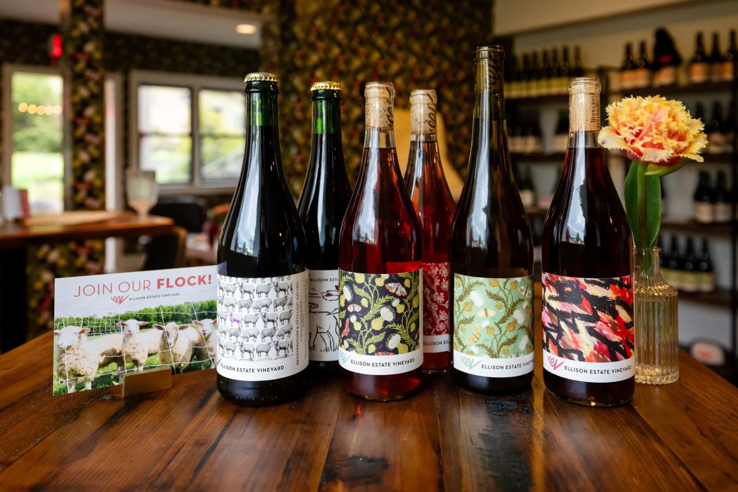 Five wine bottles with colorful labels on a wooden table, flower in a vase nearby.