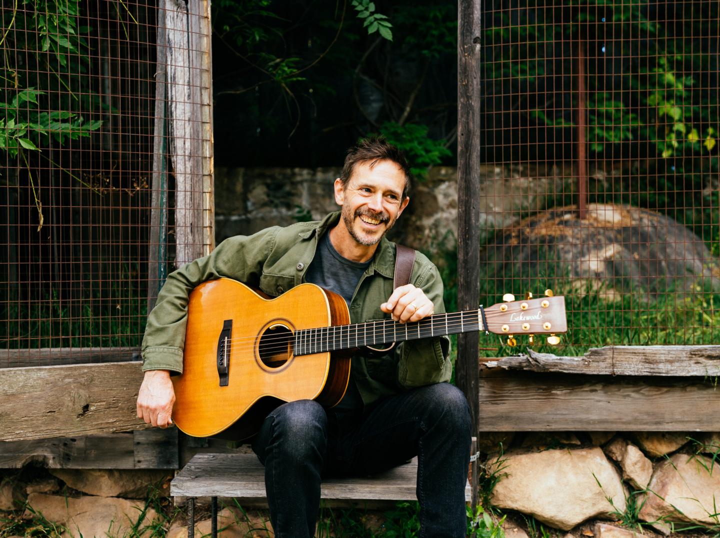 Man sitting with guitar, smiling outdoors near a wooden fence.
