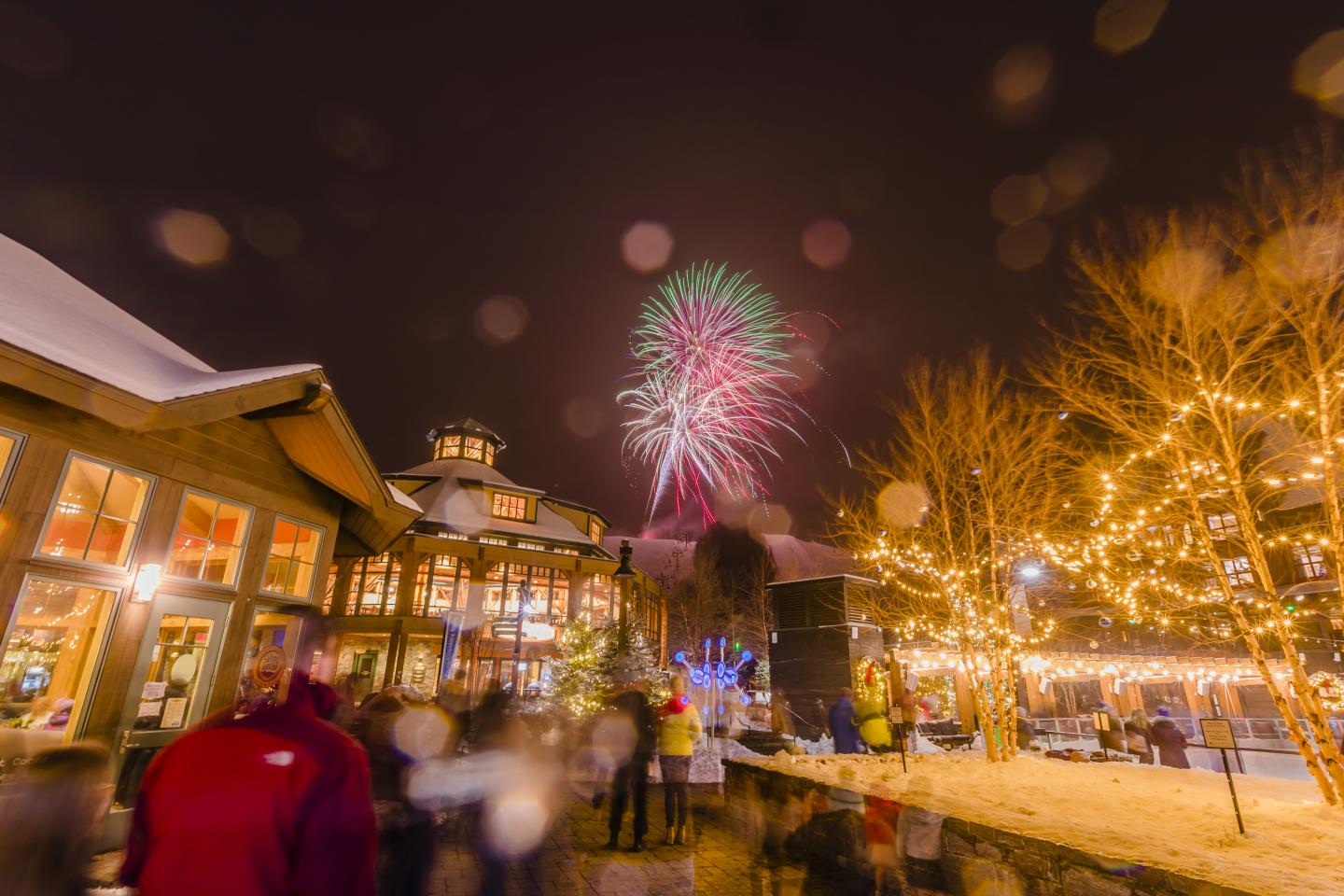 Fireworks light up a festive, winter night scene with lit buildings and trees.