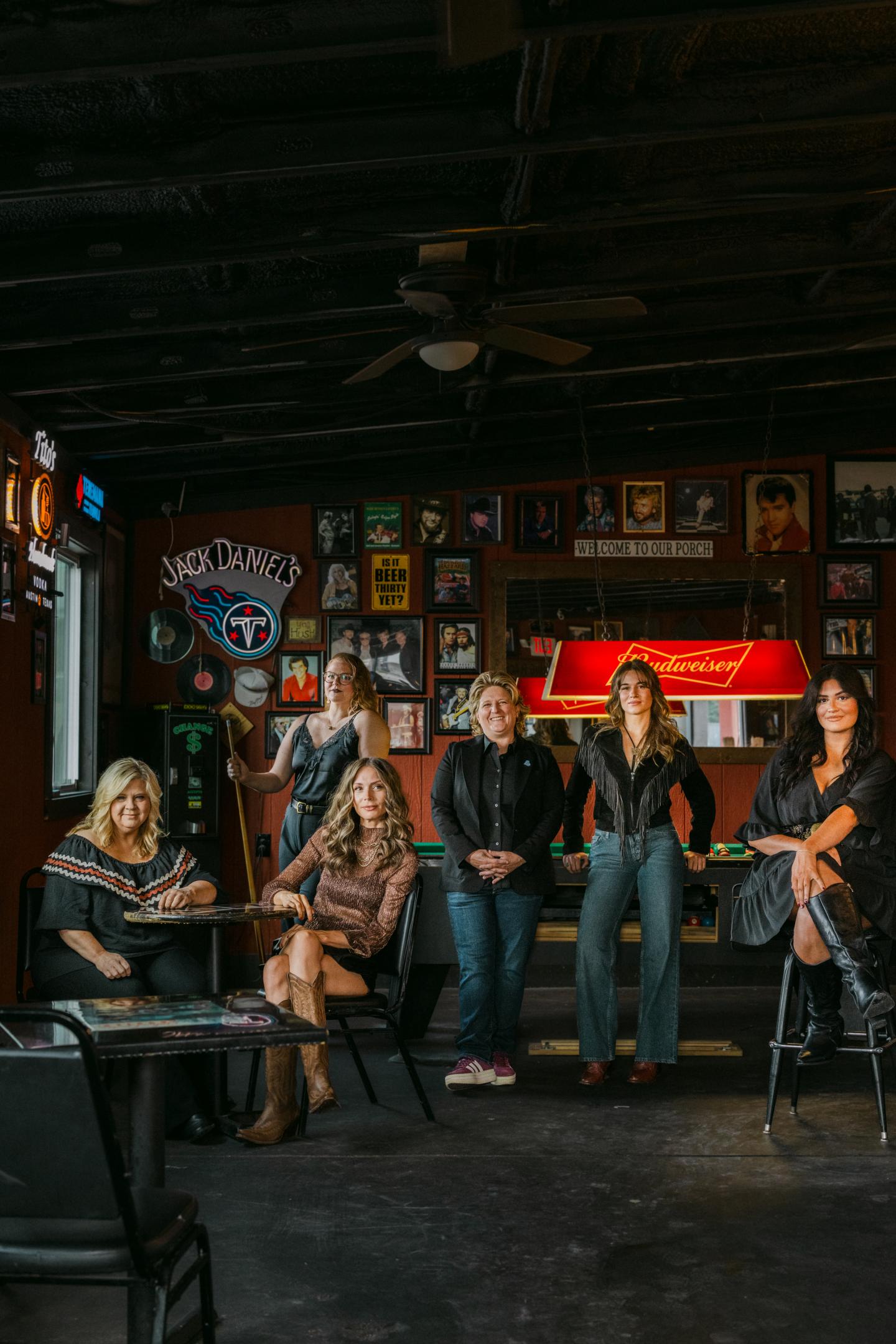 Five women in a bar, casually posed under dim lighting and framed photos.