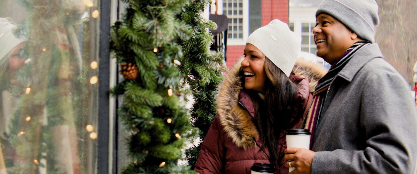 Couple in winter attire smiles while window shopping with coffee.