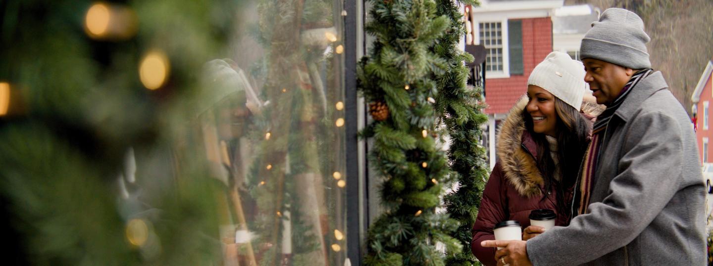 Couple window shopping, smiling, holding coffee, in winter attire.