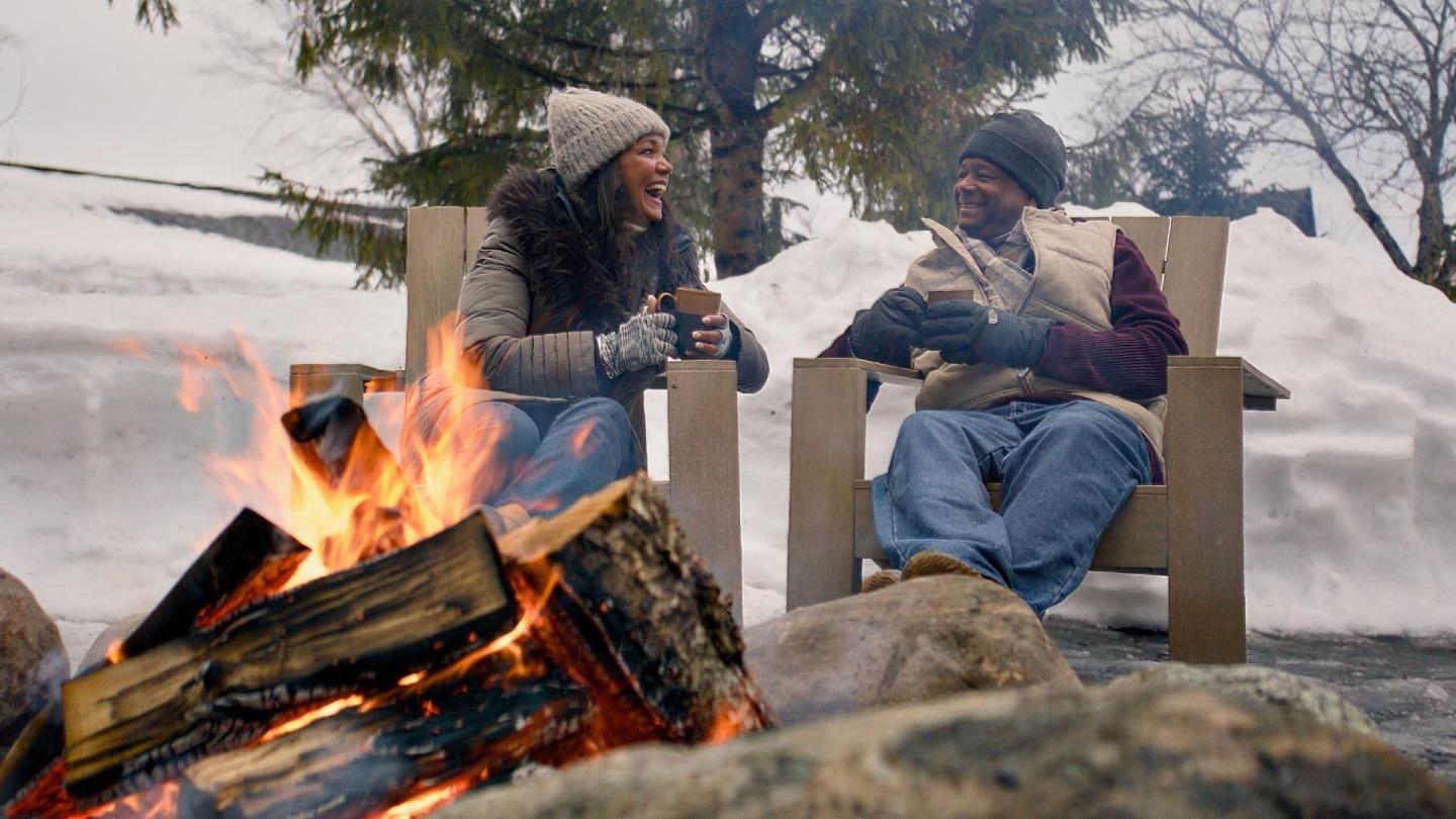 Two people laughing by a campfire in a snowy setting, wearing winter clothes.