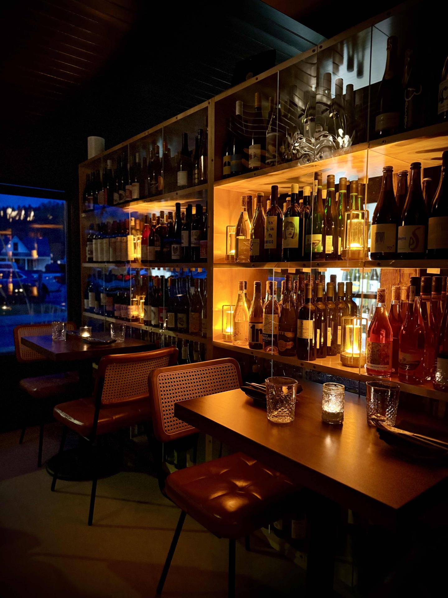 Cozy bar with dim lighting, shelves of bottles, and two small tables.