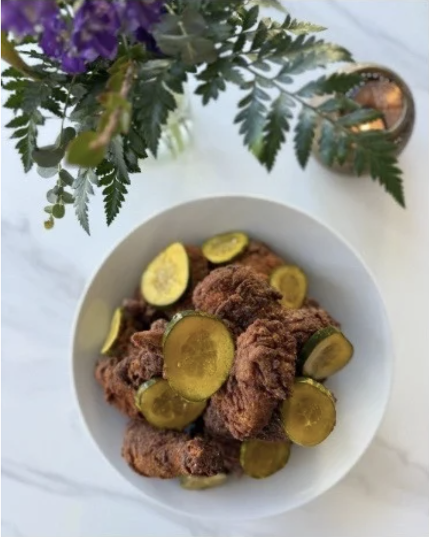 Fried chicken topped with pickles in a bowl, next to a vase of purple flowers.