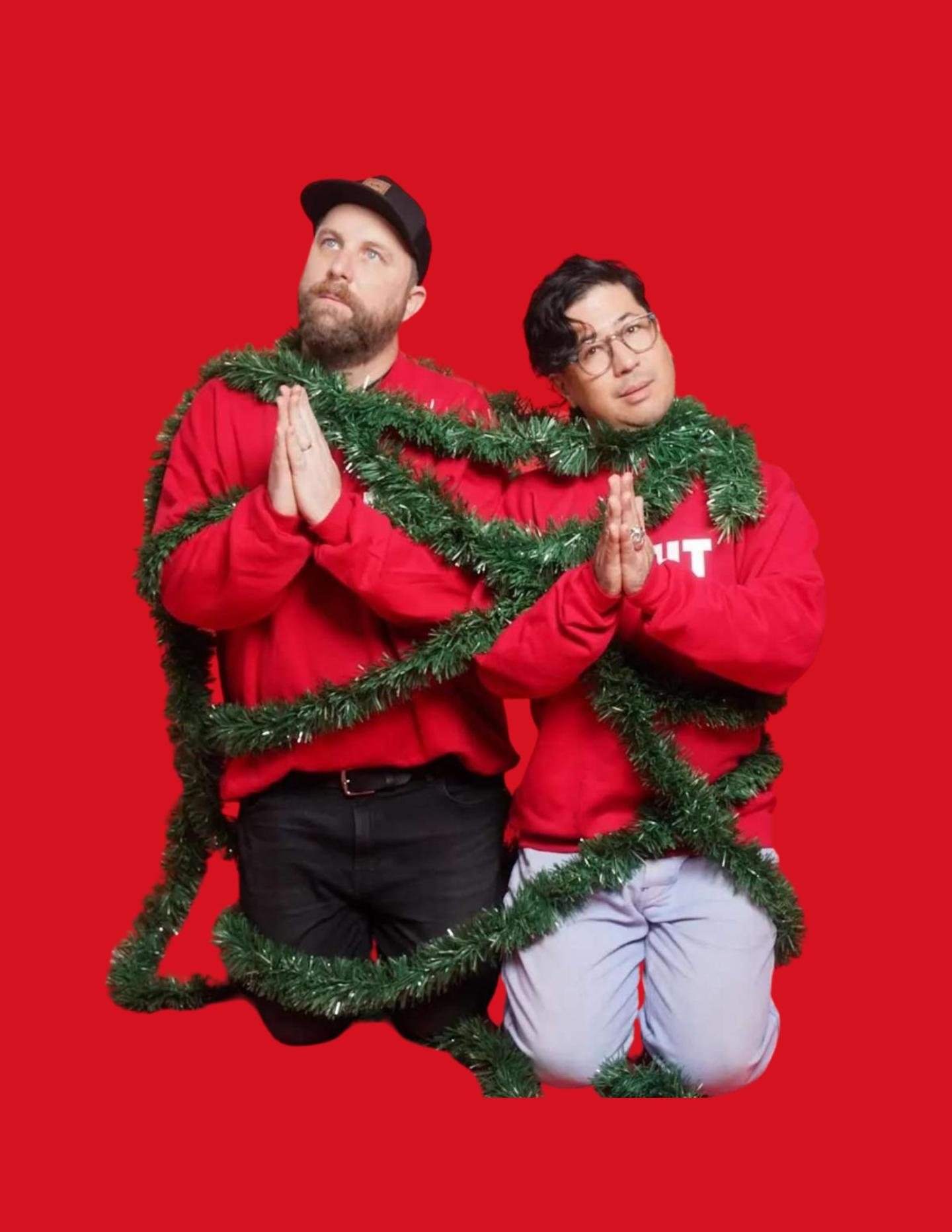 Two people kneeling, wrapped in green garland, wearing red sweaters.