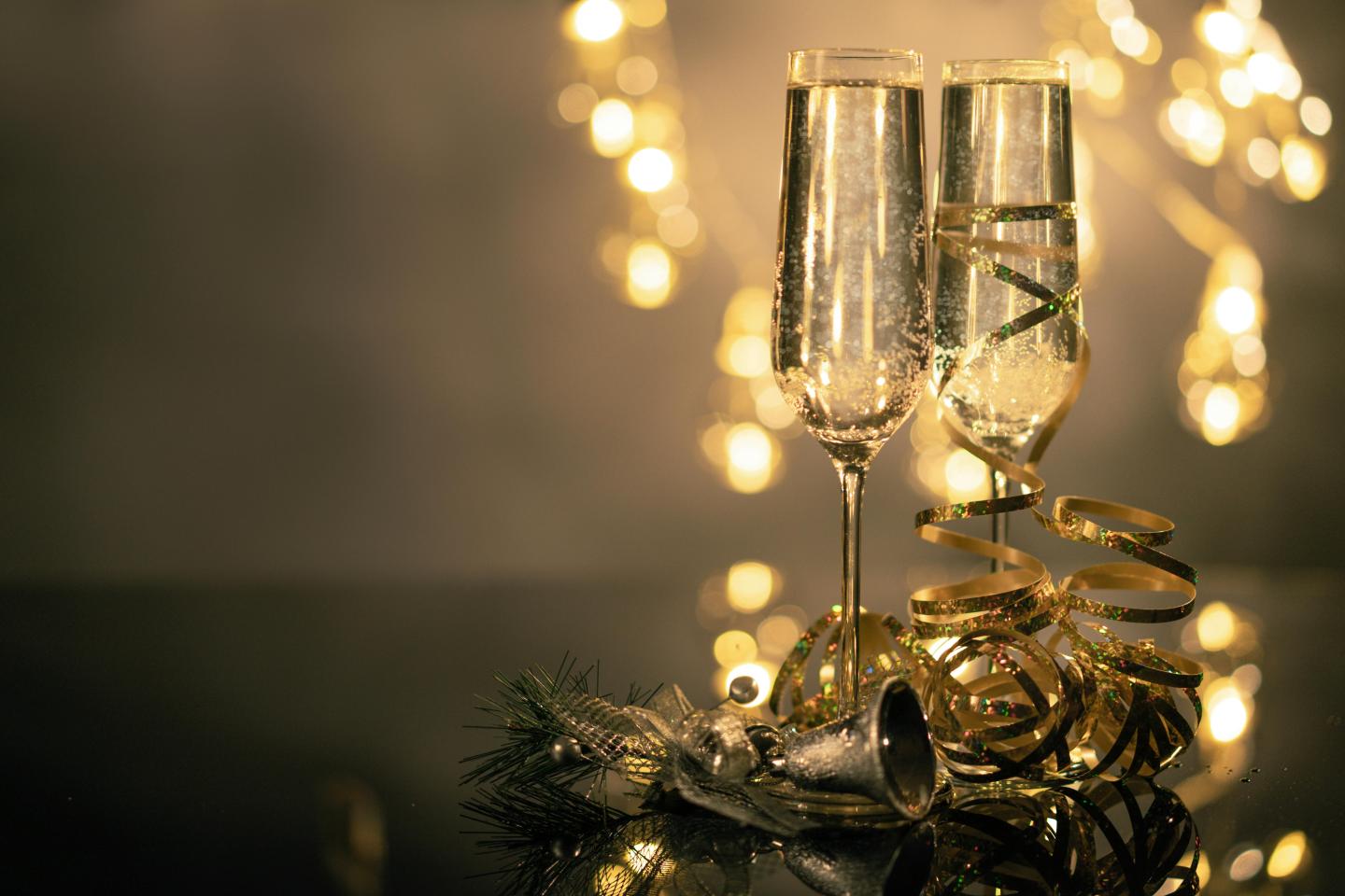 Two champagne flutes with gold ribbons, festive lights in background.