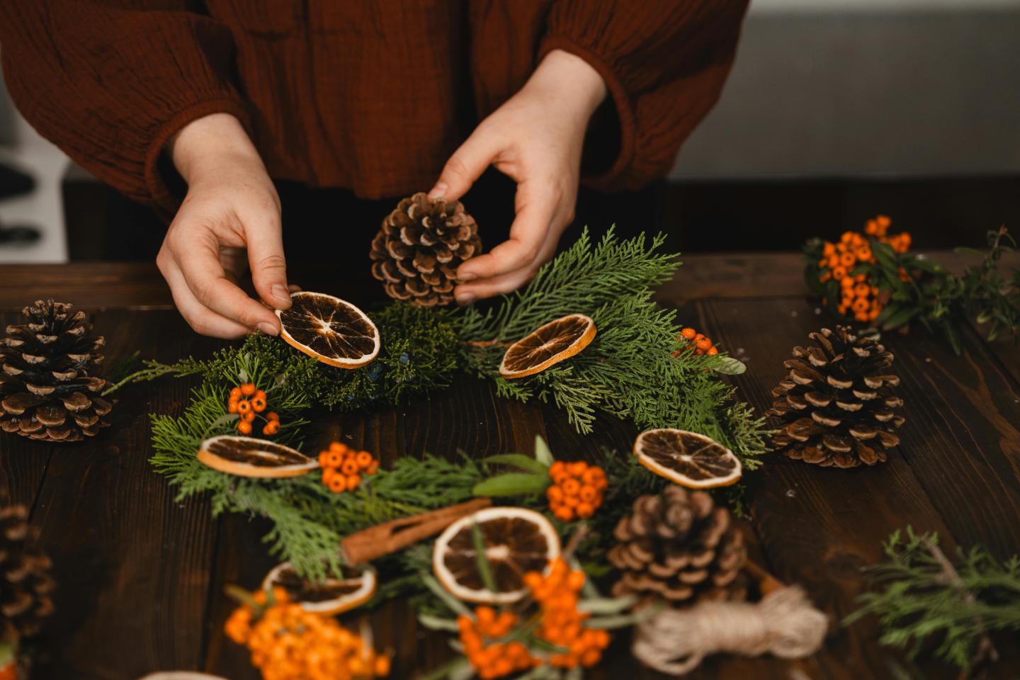 Hands decorating a wreath with pinecones, dried oranges, and orange flowers.