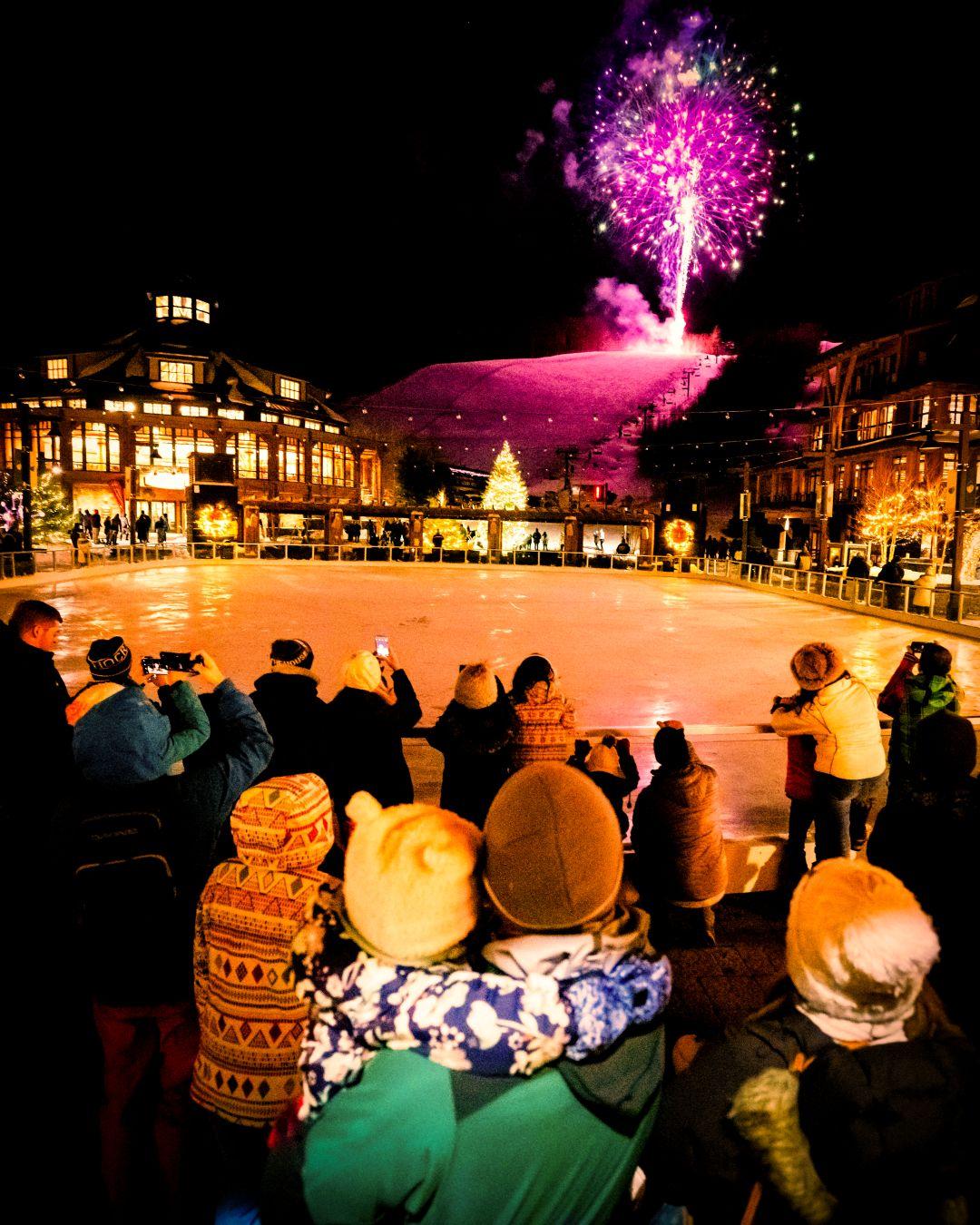 Crowd watching fireworks over an ice rink at night, surrounded by buildings.
