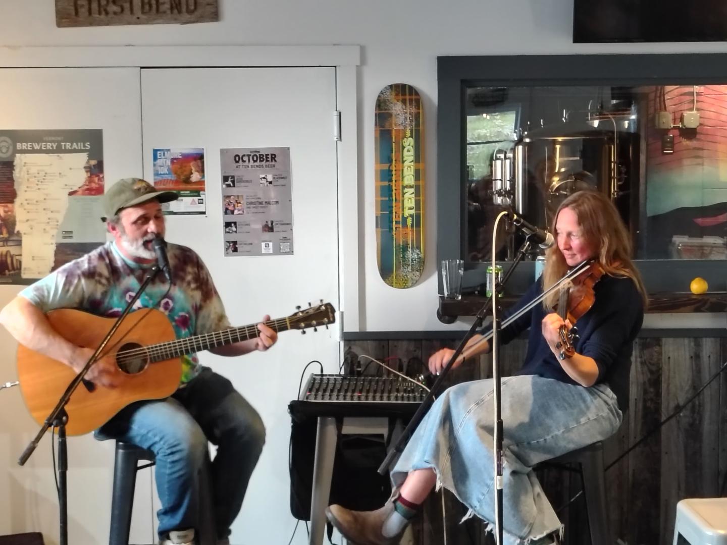 Musicians playing guitar and fiddle in a cozy room with posters on the wall.