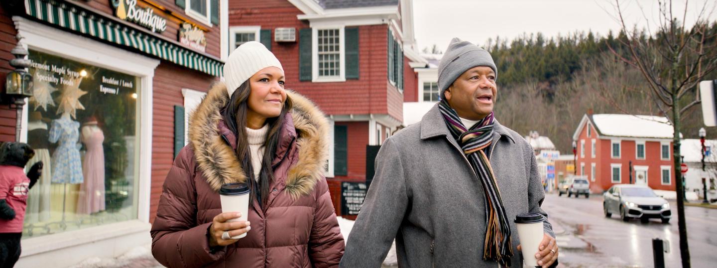 Couple walking with coffee cups in a snowy town, dressed warmly.