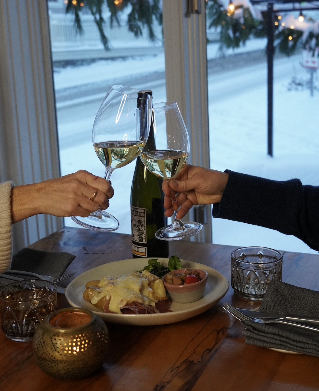 Two people clinking wine glasses over a brunch plate, snowy view outside.