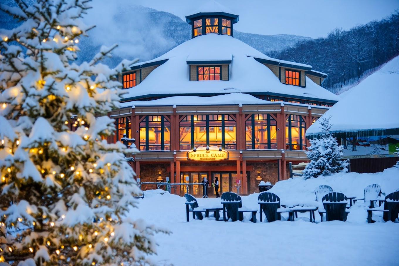 Snowy lodge with glowing windows, surrounded by mountains and a lit Christmas tree.