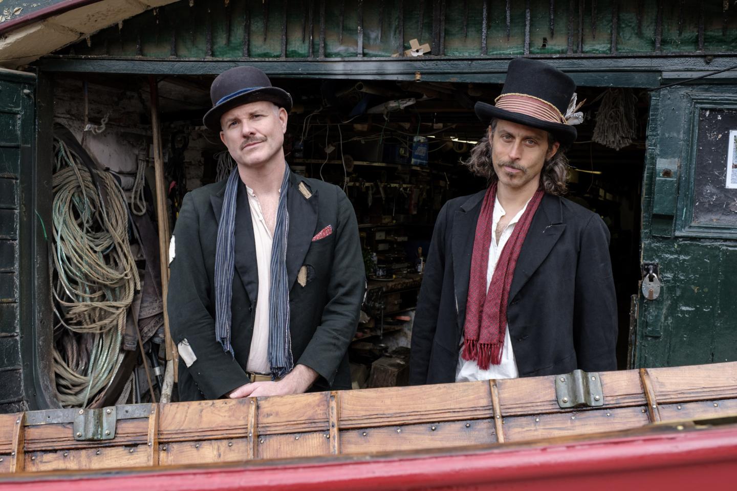 Two men in vintage attire with hats, standing in a rustic shop entrance.