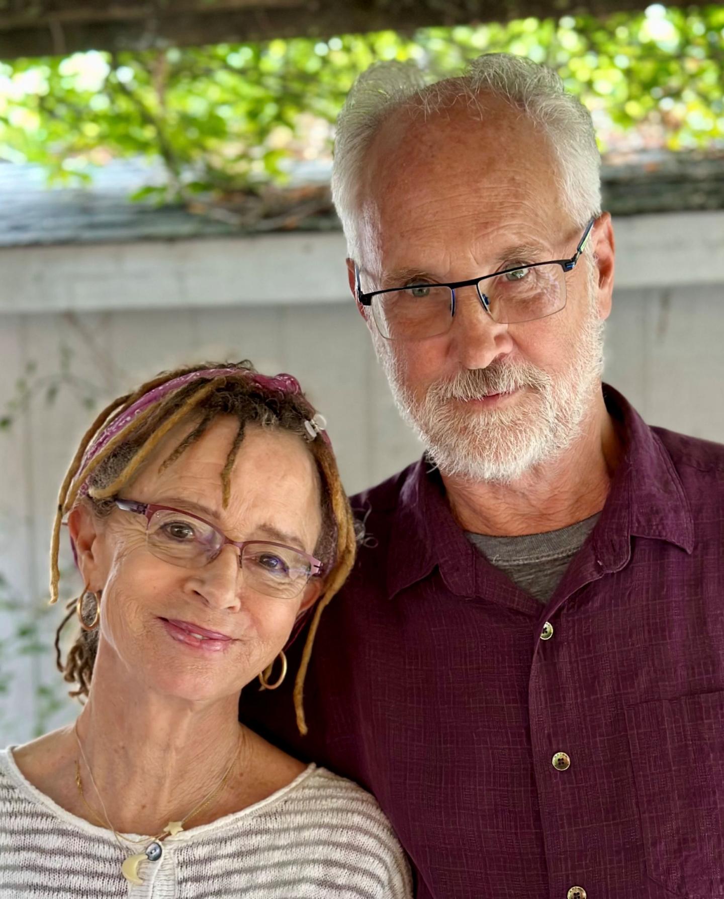 Older couple smiling, standing close, outdoors with greenery in background.