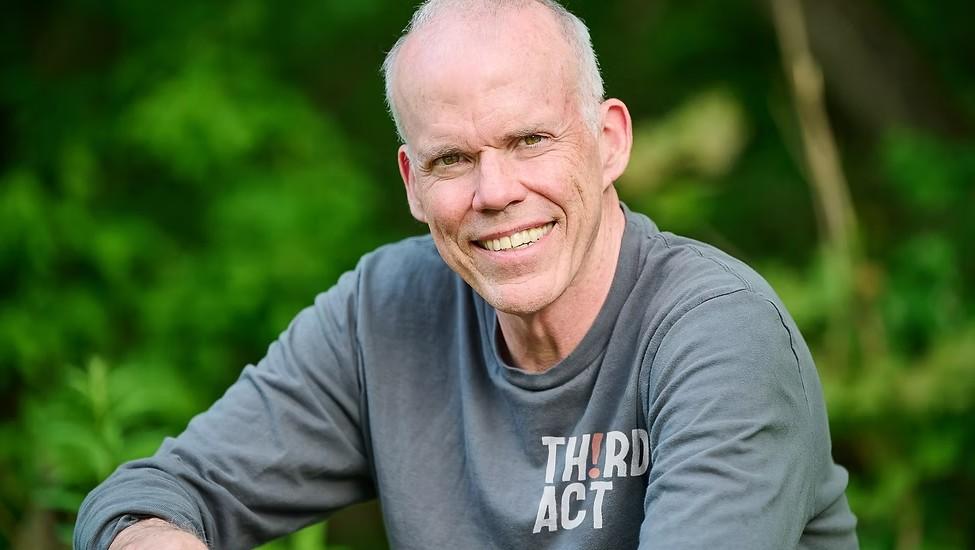 Smiling man in a grey shirt outdoors with greenery behind.