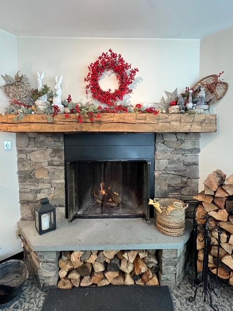 Stone fireplace with red wreath and festive decorations. Logs stacked nearby.