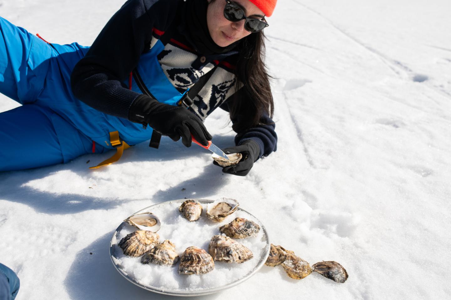 Person in snow with oysters, shucking one with a knife, wearing blue gear and red hat.