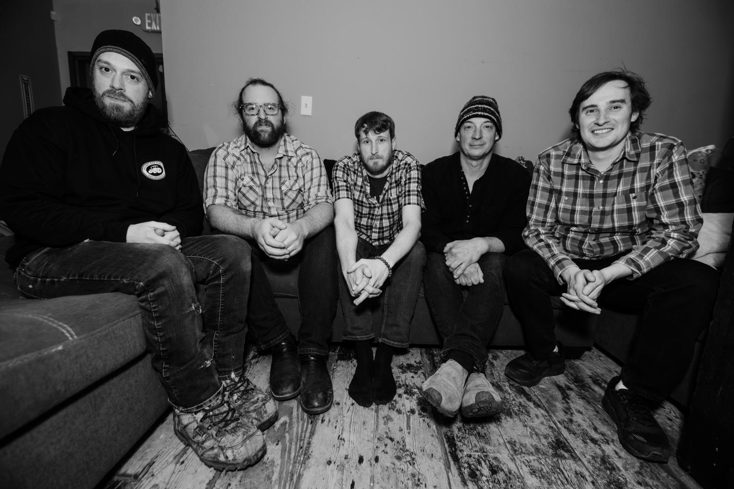 Five men sitting on a couch, smiling, in a black and white photo.
