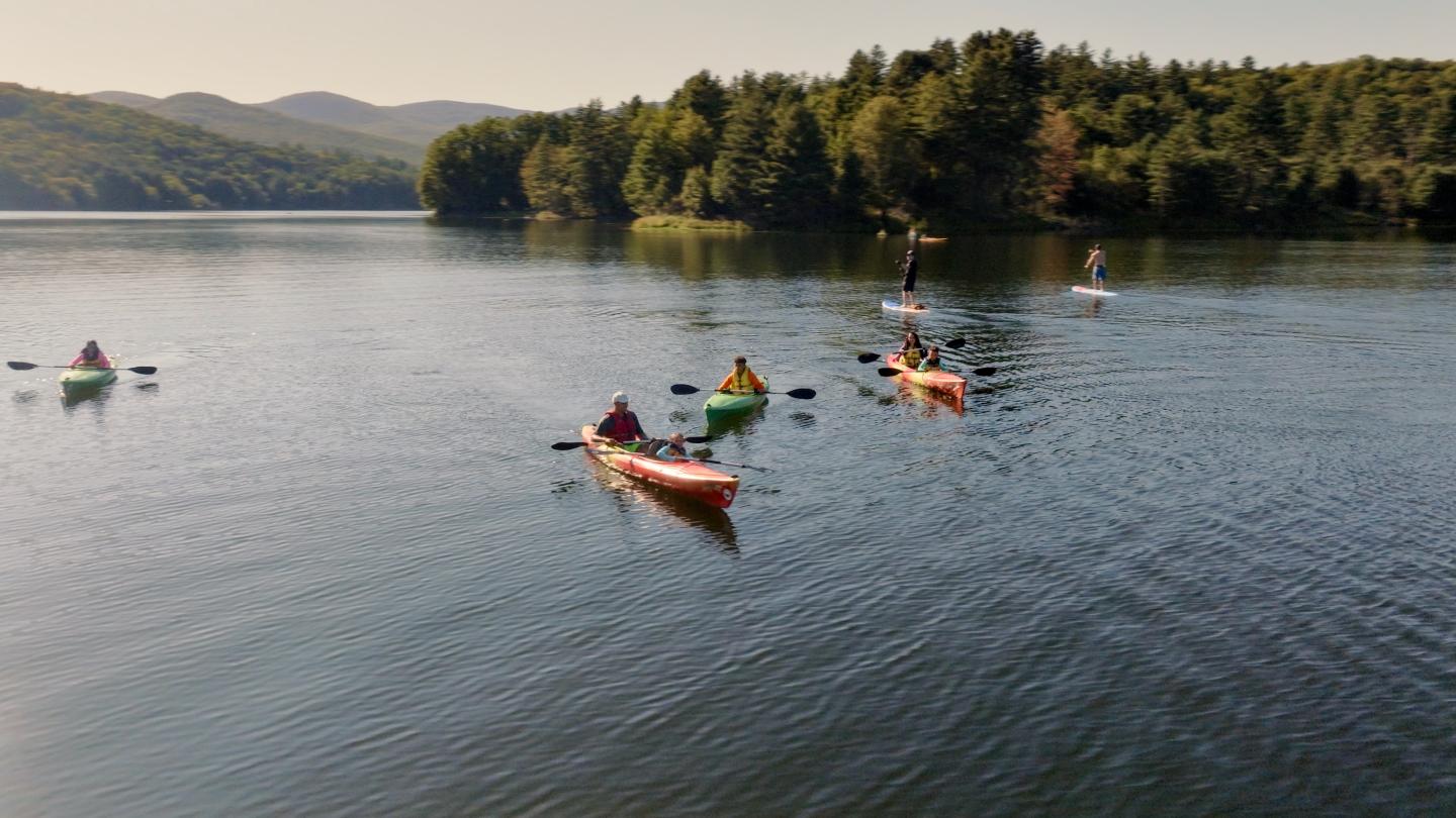 Kayakers paddle on a calm lake surrounded by trees and hills.
