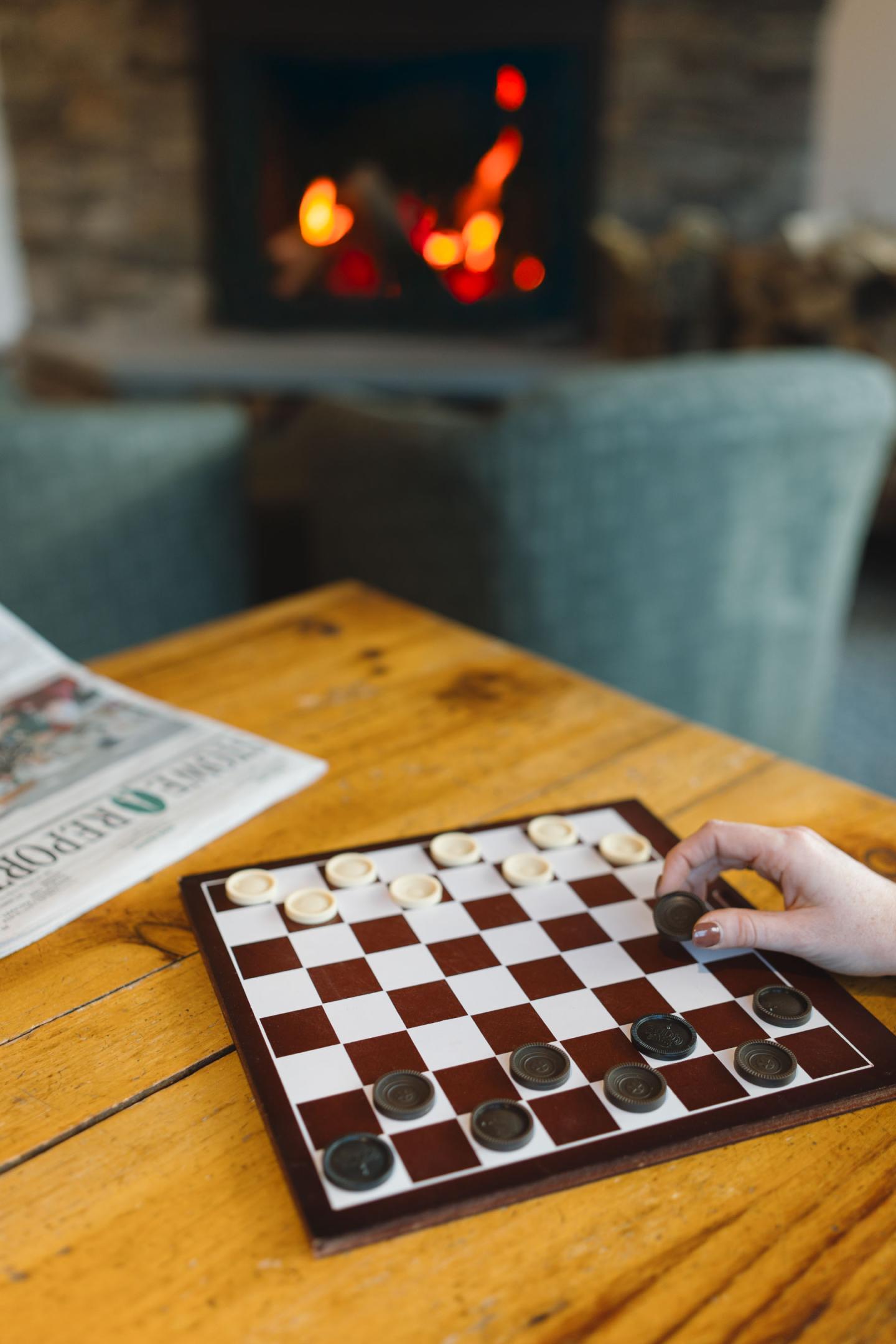 Checkerboard on a table, hand moving a piece, with a fireplace in the background.