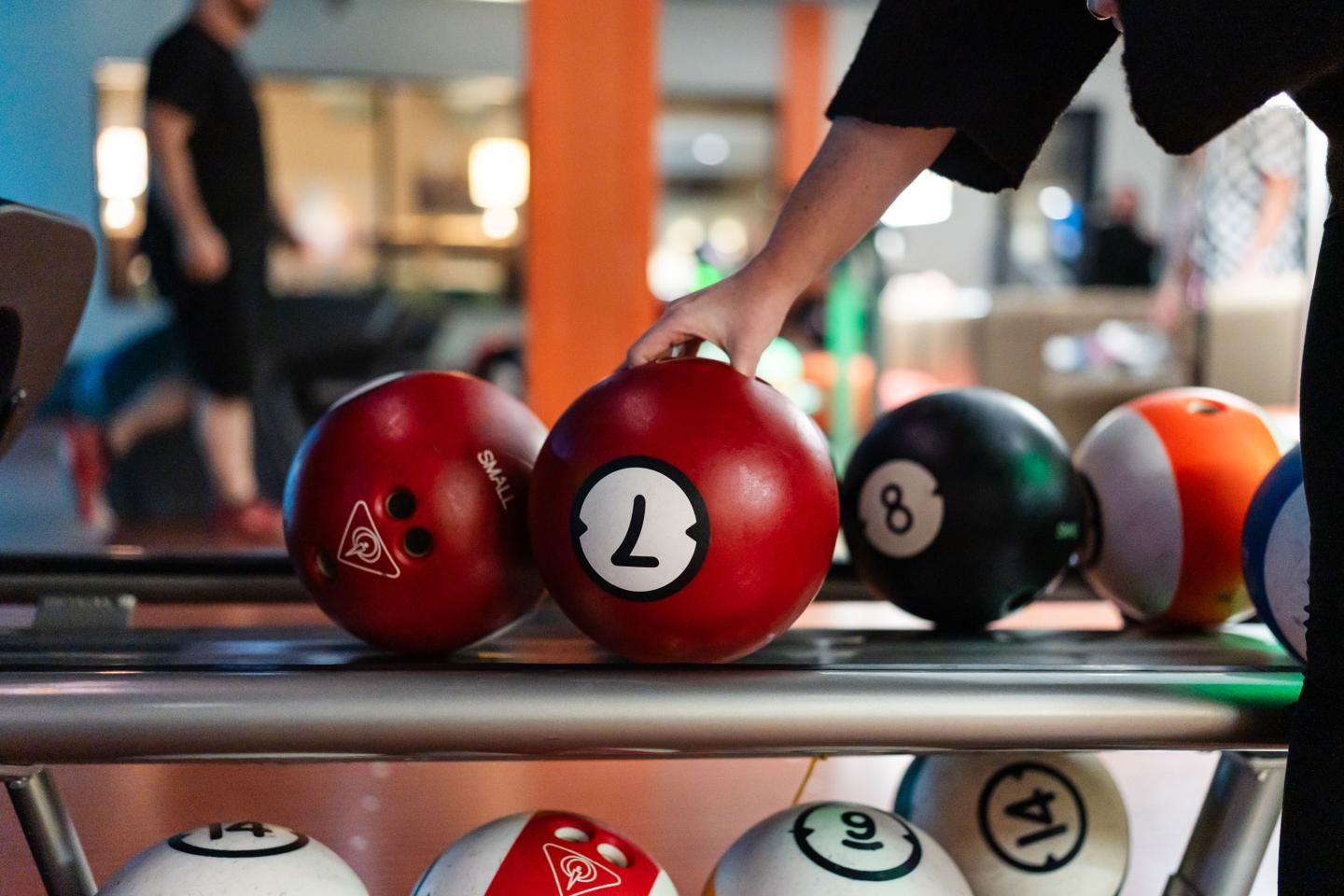 Bowling balls on a rack, hand reaching for a red one with a "7" in a bowling alley.