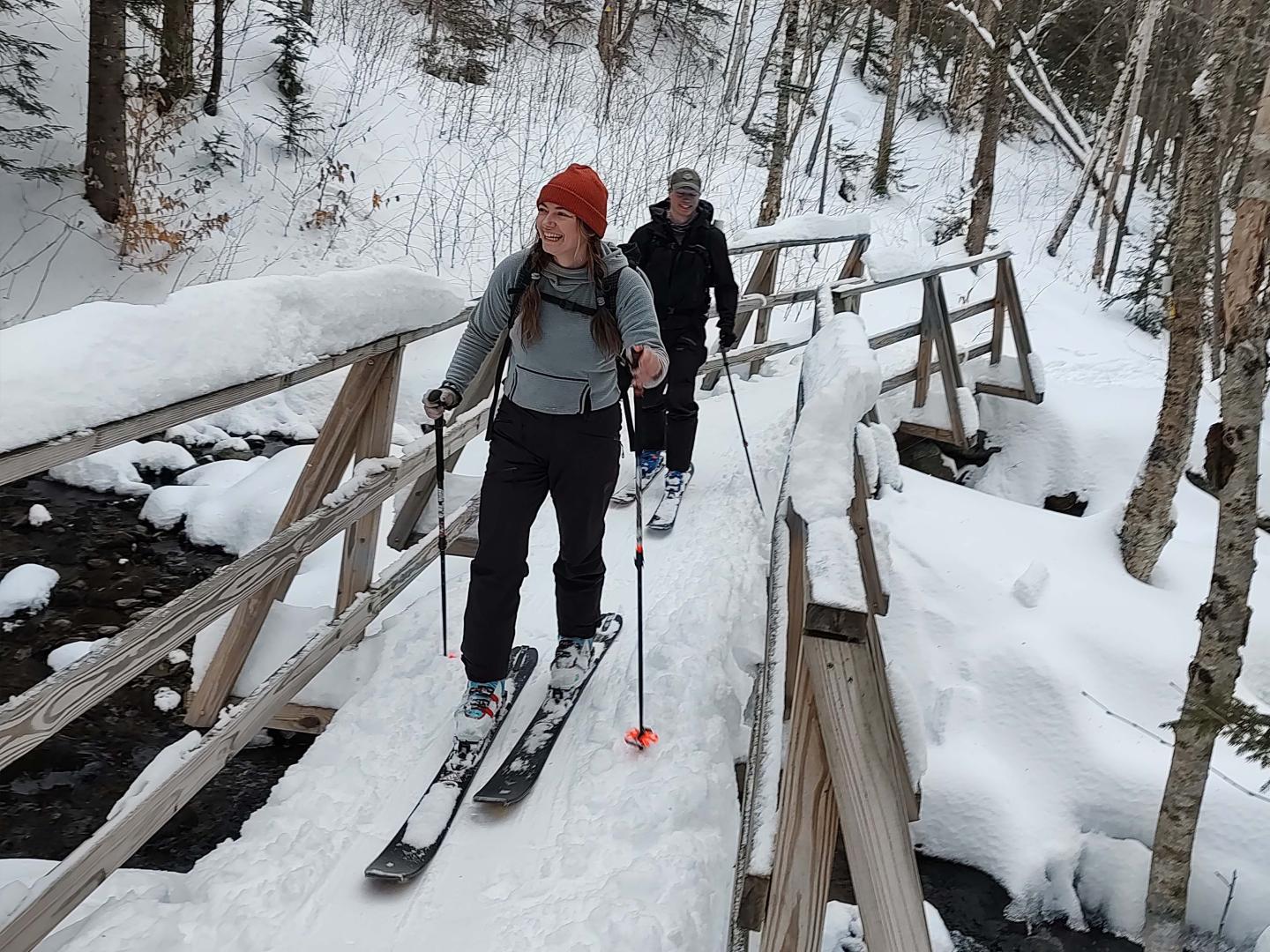 Two people cross a snowy bridge on skis in a forest.