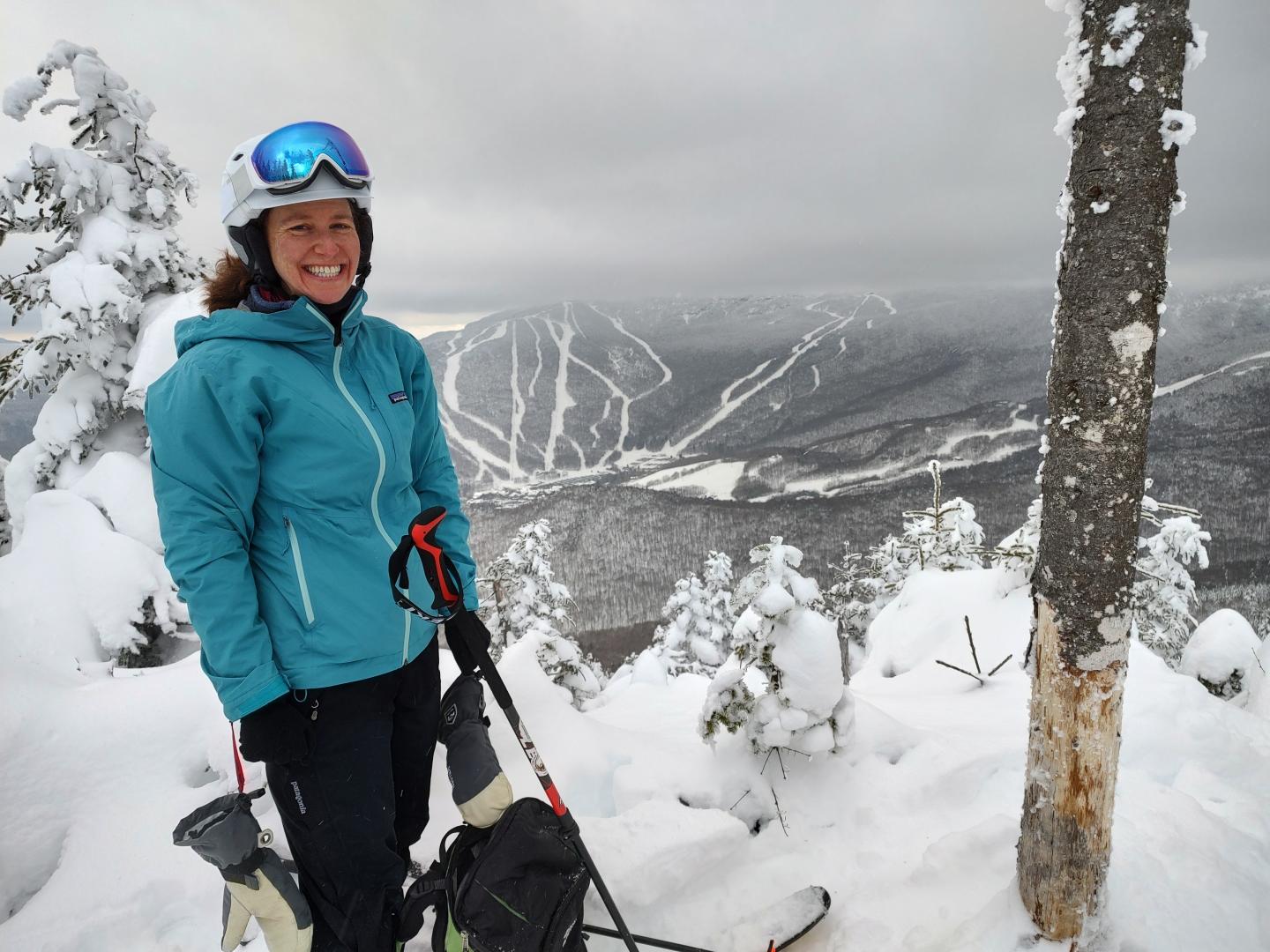 Smiling skier in blue jacket on snowy mountain with cloudy sky.